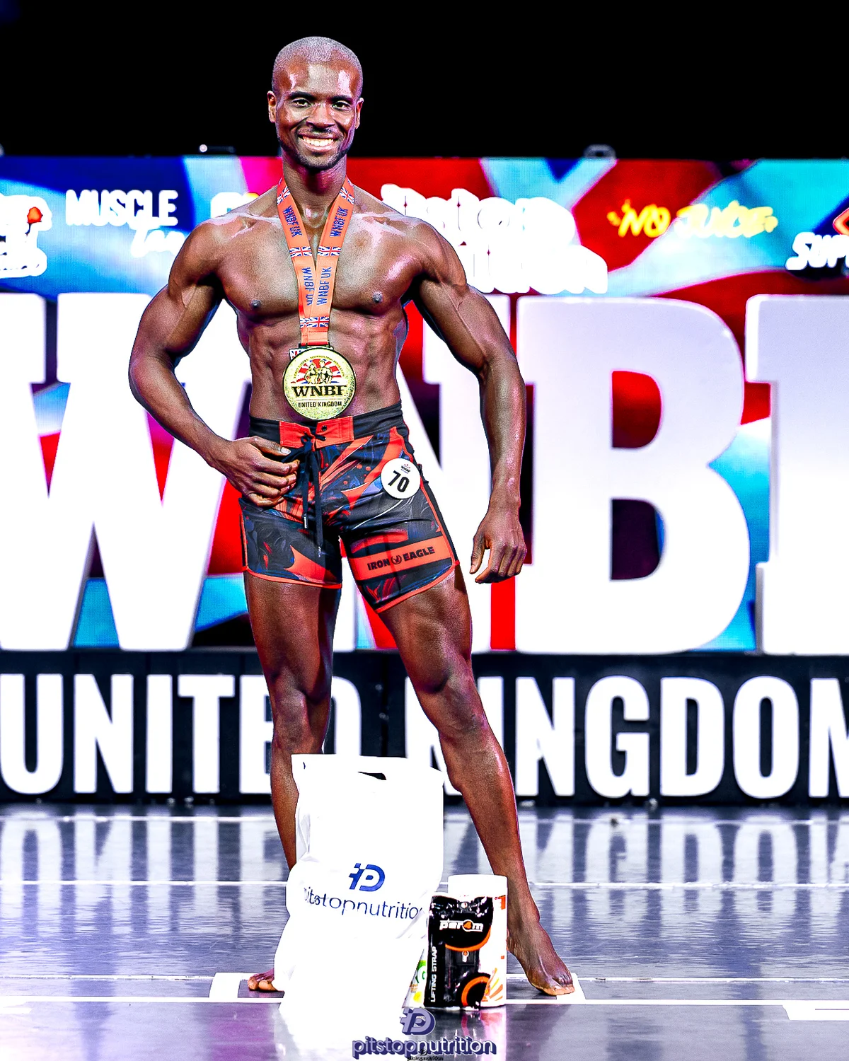Femi celebrating a WNBF Men's Physique Short title win on stage with his medal