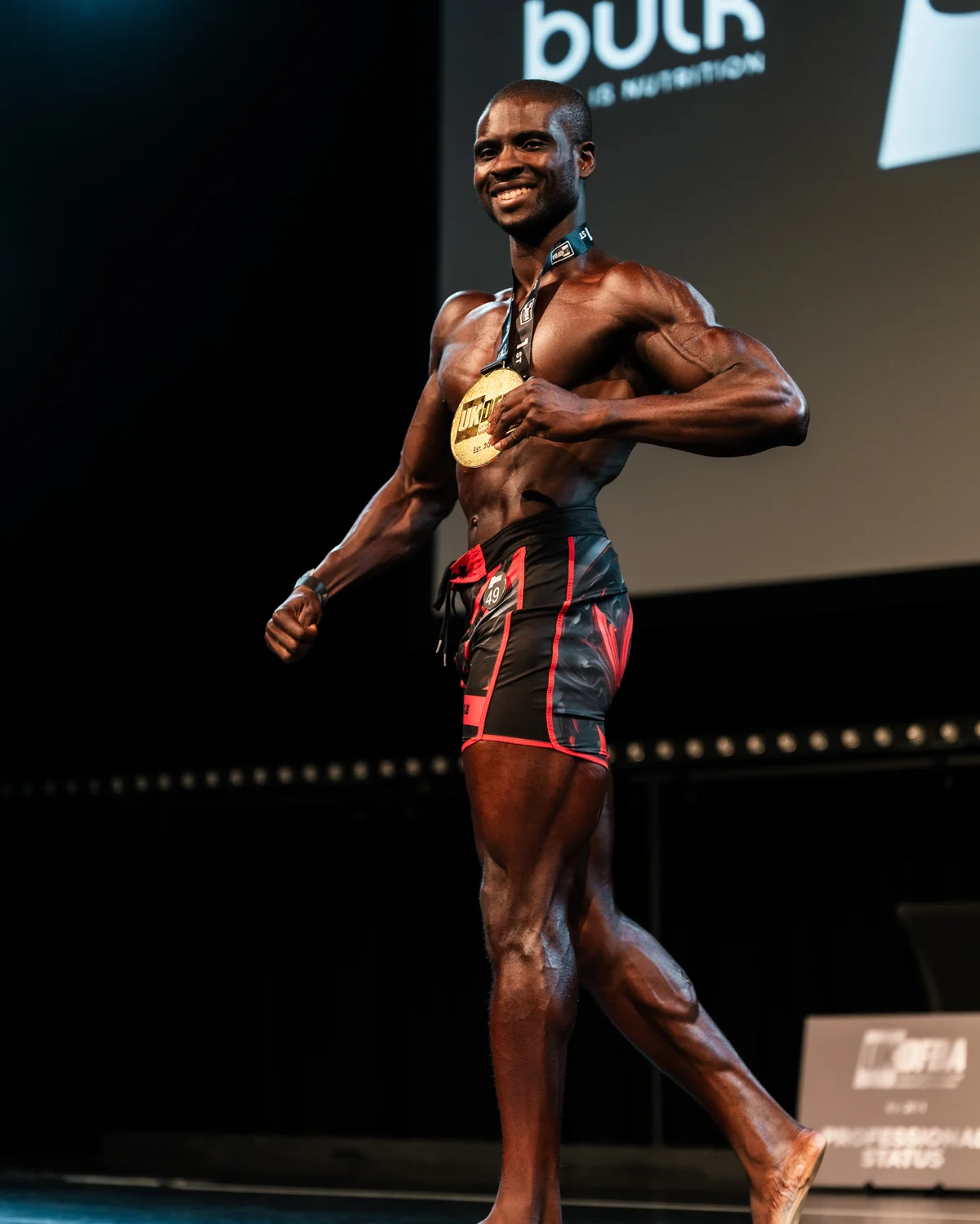 Femi celebrating a UKDFBA Men's Physique Short title win on stage with his medal